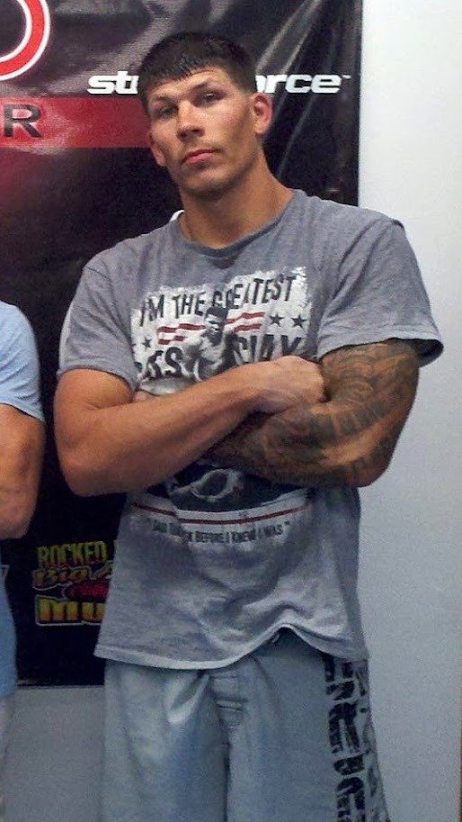 Dustin Baker in his fighting years, arms crossed, wearing a gray Muhammad Ali t-shirt